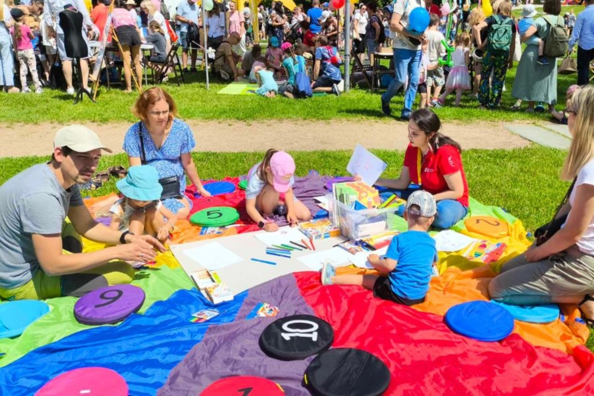Grupa dzieci i dorosłych siedzi na kolorowym kocu na trawie podczas pikniku. Dzieci rysują kredkami na dużych kartkach, a wokół rozłożone są kolorowe poduszki z liczbami. W tle widoczni inni uczestnicy wydarzenia z balonami i namiotami.